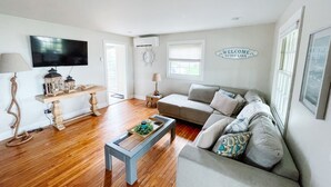 A living room with a sofa, coffee table, and TV. There is a door leading outside, a window, and a wall-mounted welcome sign. - Private Waterfront Belle Point Shores Cottages (Leamington)