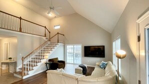 Ein geräumiger Wohnbereich mit einem weißen, gebogenen Sofa, einer Holztreppe und einem Deckenventilator.