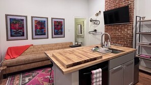 A compact kitchen with a wooden countertop, a built-in sink, and a black dishwasher. There is a sofa with a red blanket and a colorful rug in the adjacent living area. - Welcome to the JazzNest!   (New Orleans)
