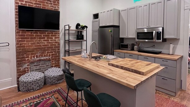 A modern kitchen with a brick accent wall, a flat-screen TV, grey cabinets, a refrigerator, a microwave, and a wooden island with bar stools. - Welcome to the JazzNest!   (New Orleans)