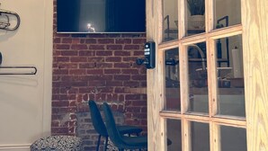 A room with a brick wall, a mounted TV, a wooden door, and two black bar stools. - Welcome to the JazzNest!   (New Orleans)