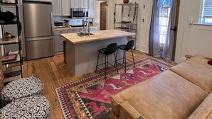 A modern kitchen with white cabinets, a stainless steel refrigerator, a microwave, and a built-in oven. There is a wooden dining table with two chairs and a rug with a geometric pattern. - Welcome to the JazzNest!   (New Orleans)