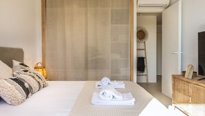 Een net opgemaakt bed met opgerolde handdoeken, een rieten dressoir, een televisie en een zondecoratie aan de muur.