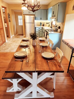 Comedor con una mesa de madera dispuesta para seis personas, una araña y una cocina al fondo.