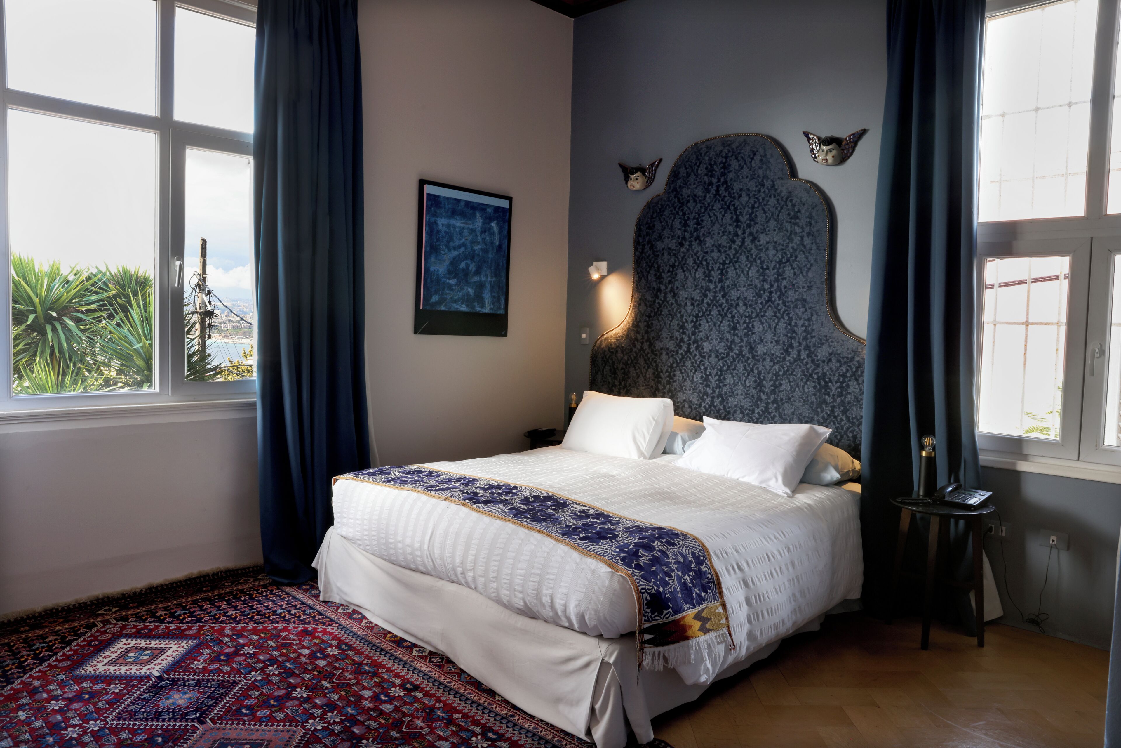 Ein Schlafzimmer mit einem großen Bett, einem Kopfteil, einem Nachttisch, einem Schreibtisch, einem Fenster mit Vorhängen und Blick ins Grüne.