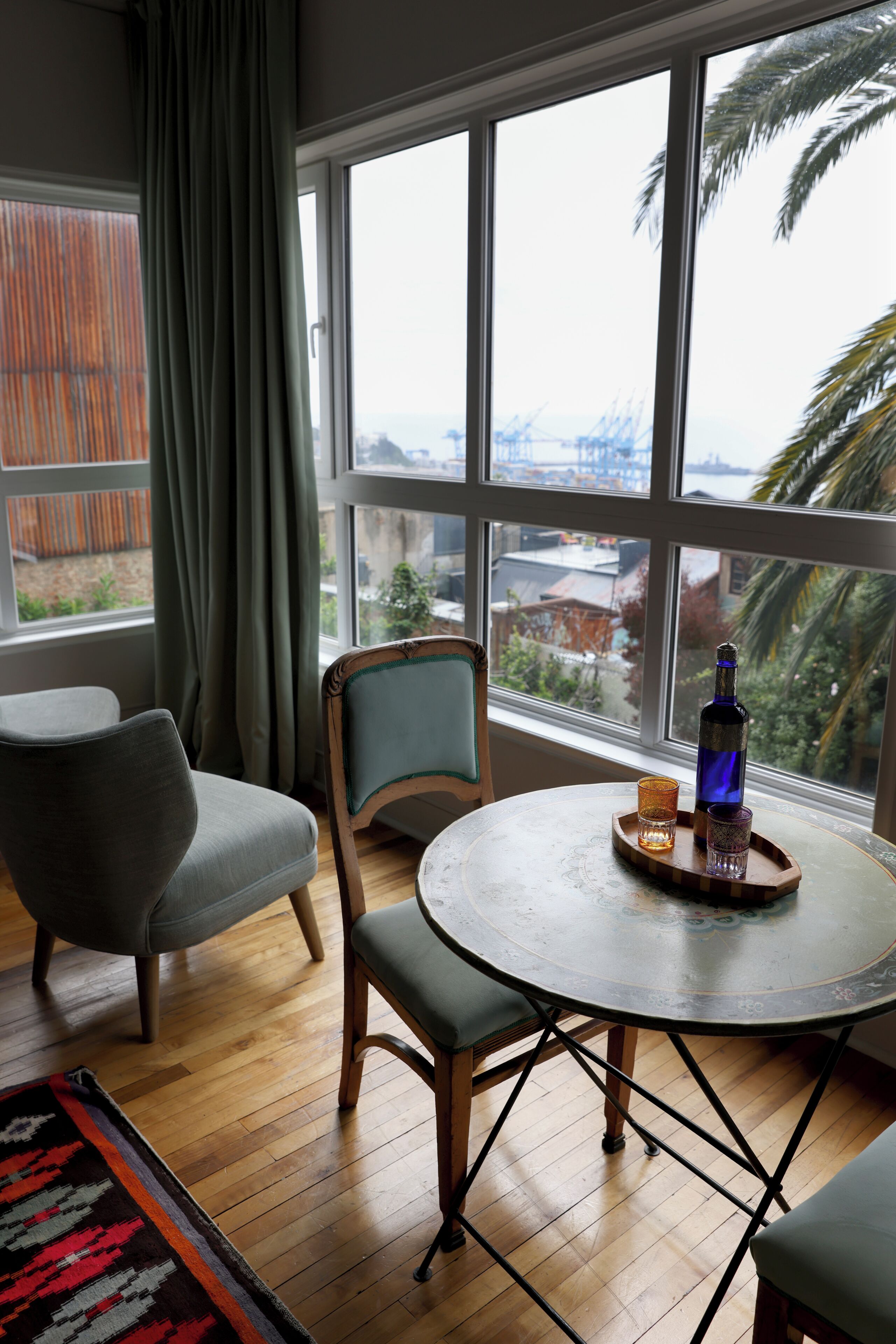 Una habitación con un ventanal grande, una mesa redonda con una botella y vasos, una silla y una alfombra.
