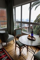 Una habitación con un ventanal grande, una mesa redonda con una botella y vasos, una silla y una alfombra.