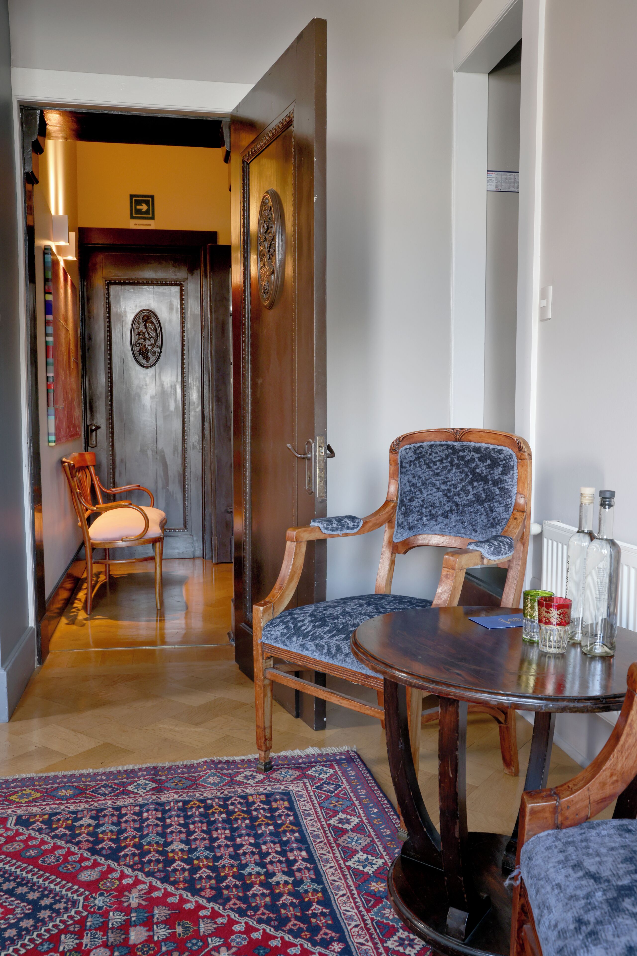 Una habitación con puerta de madera, un sillón tapizado en azul, una mesita redonda y una alfombra estampada.