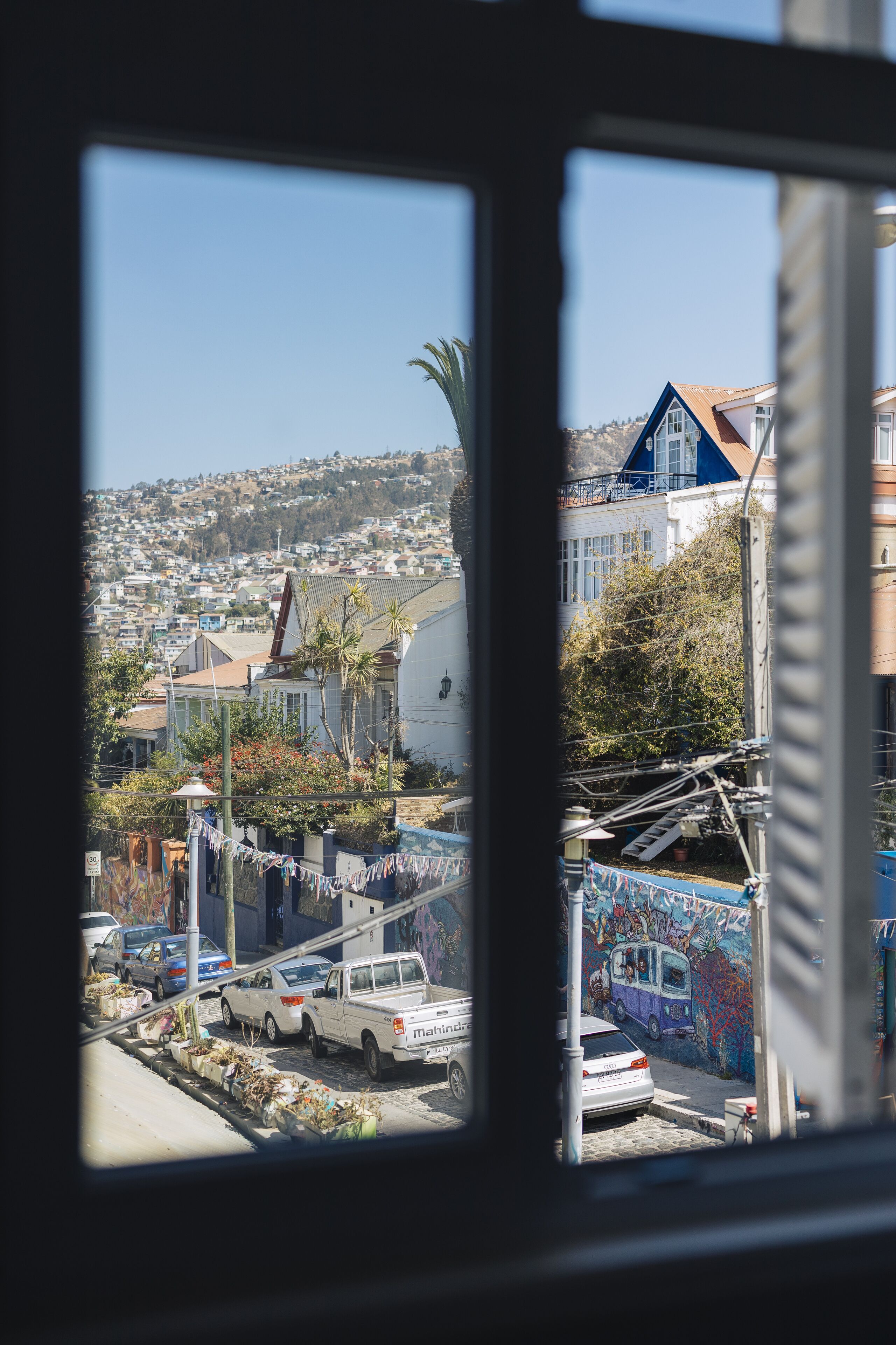 Vista a través de una ventana que muestra una zona residencial con coches aparcados, una furgoneta blanca y grafitis de colores en una pared.