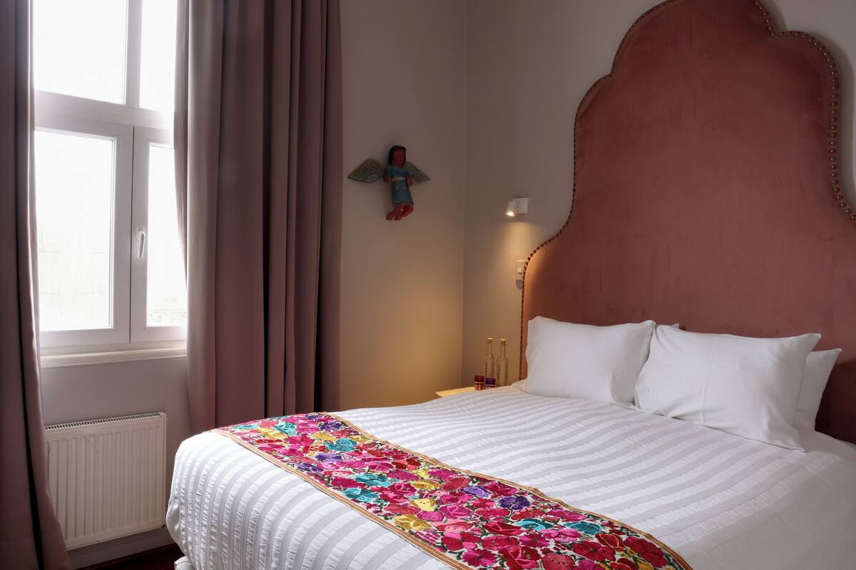 Habitación de hotel con una cama grande, una ventana con cortinas, una mesita de noche con botellas y una decoración de ángel en la pared.