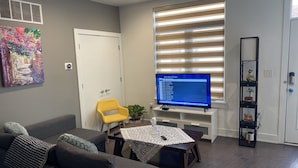 A modern living room with a grey sofa, a yellow chair, a coffee table with a lace tablecloth, a TV on a stand, and a bookshelf with decorative items.