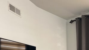 A room with a flat-screen TV mounted on the wall, a power outlet, a mirror, and a bed with a green blanket.