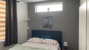 A bedroom with a bed, a nightstand, a lamp, a window with curtains, and a painting on the wall.