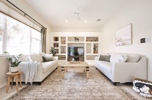 A modern living room with two white sofas, a wooden coffee table, and a large TV. There are shelves with decorative items and a ceiling fan. - The happy family Hideaway (Port Charlotte)
