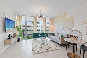 Uma sala de estar moderna com sofá, mesa de centro e cadeiras, uma televisão e uma janela grande com vista para edifícios.