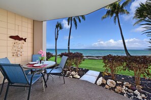 A covered outdoor seating area with a table and chairs, overlooking a beach with palm trees and the ocean.