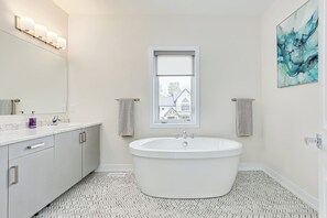 Ett modernt badrum med ett fristående badkar, en stor spegel och ett fönster med utsikt över hus.