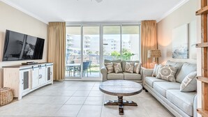 Ein geräumiges Wohnzimmer mit einem Flachbildfernseher, einem runden Couchtisch, einer Couch und einem Balkon mit Blick auf Gebäude und Grünflächen.