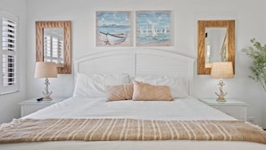 Een slaapkamer met een wit bed, twee schilderijen erboven en twee houten spiegels aan weerszijden.