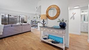 Een moderne woonkamer met een grijze bank, houten vloeren, een ronde spiegel en een console met een bord waarop 'BEACH HOUSE' staat.
