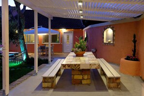 An outdoor patio with a table and benches, surrounded by a garden and a building with a door and windows.