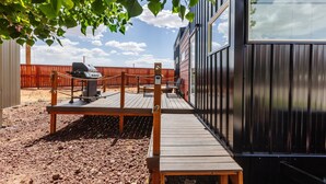 Eine Terrasse mit Grill, Holzbanken und einem eingezäunten Bereich, im Hintergrund ein Metallgebäude.