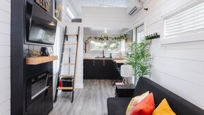 Ein kompakter Wohnraum mit einer Küche, einem schwarzen Sofa mit bunten Kissen, einem an der Wand befestigten Fernseher und einer Leiter zu einem Schlafbereich auf Podest.