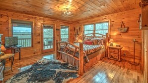 Een houten slaapkamer met een bed, een televisie, een bureau en een raam met uitzicht op een bebost gebied.