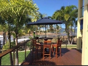 Eine Terrasse mit Tisch, Stühlen und Sonnenschirm, mit Blick auf das Meer und Palmen.