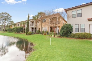 A residential area with a pond, well-maintained lawn, and houses with balconies and windows.