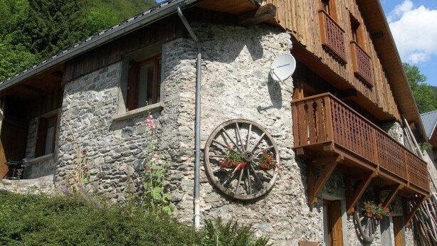 A stone building with a wooden balcony and a large wheel attached to the wall. - Gite 8 km from the access to Alpe d'Huez large domain (ALLEMOND)