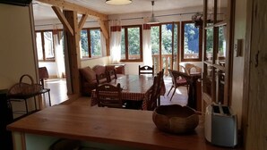 A spacious dining area with a wooden table and chairs, large windows with curtains, and a view of greenery outside. - Gite 8 km from the access to Alpe d'Huez large domain (ALLEMOND)
