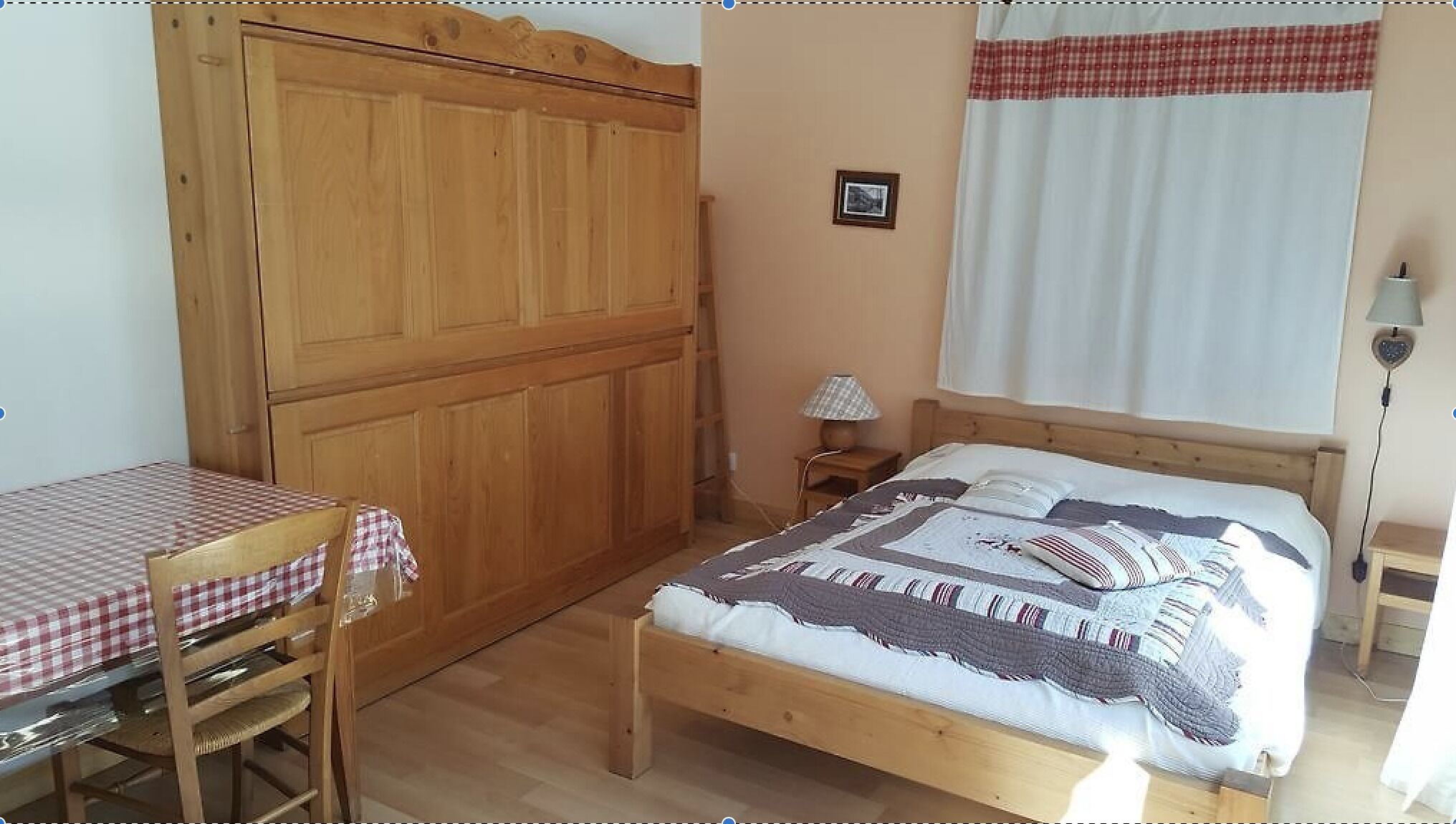 Quarto com cama de madeira, guarda-roupa de madeira, mesa de cabeceira pequena com abajur e uma mesa xadrez com uma cadeira.