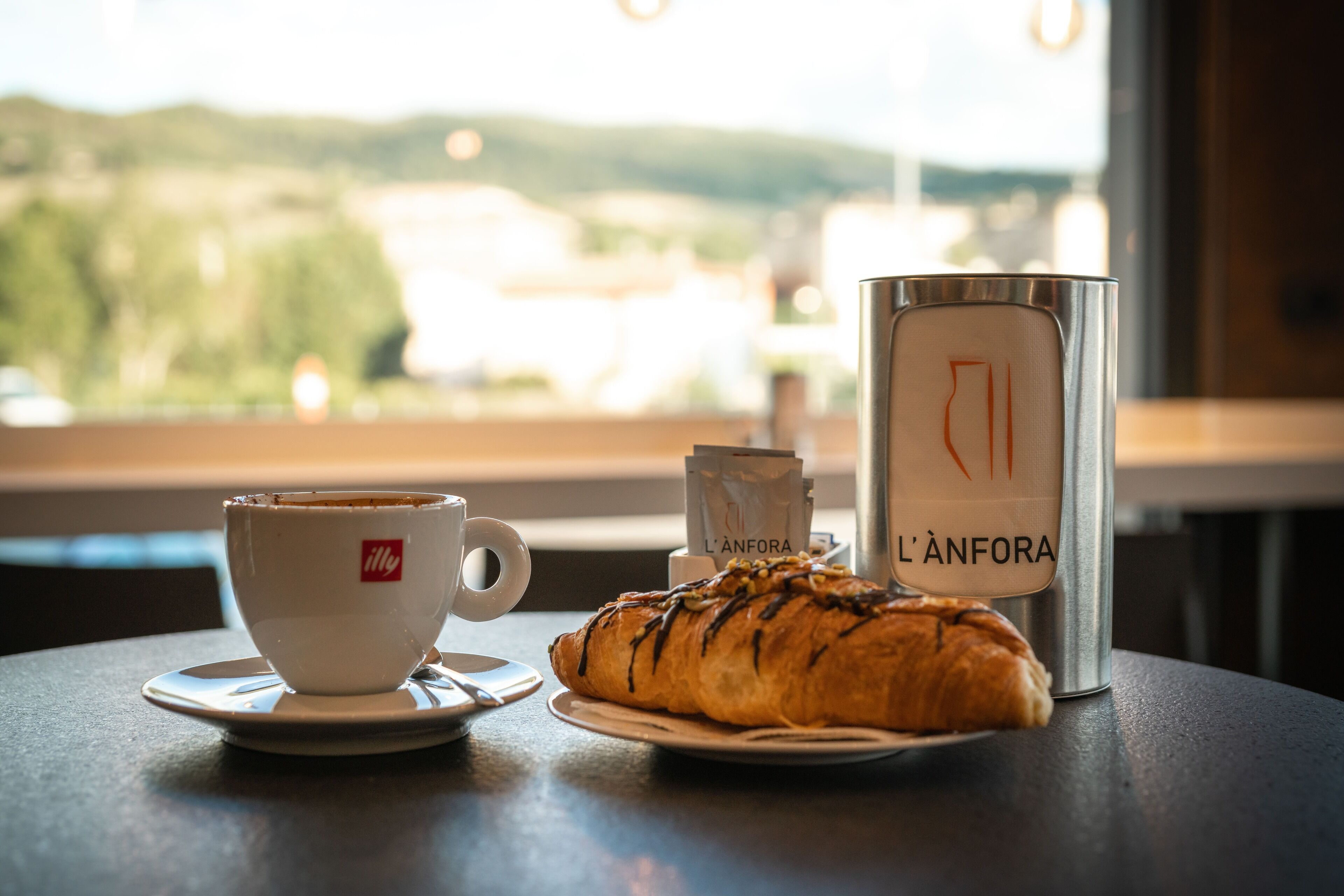A table with a croissant, a cup of coffee, and a thermos labeled "L'ANFORA" in a cafe setting with a view of a landscape.