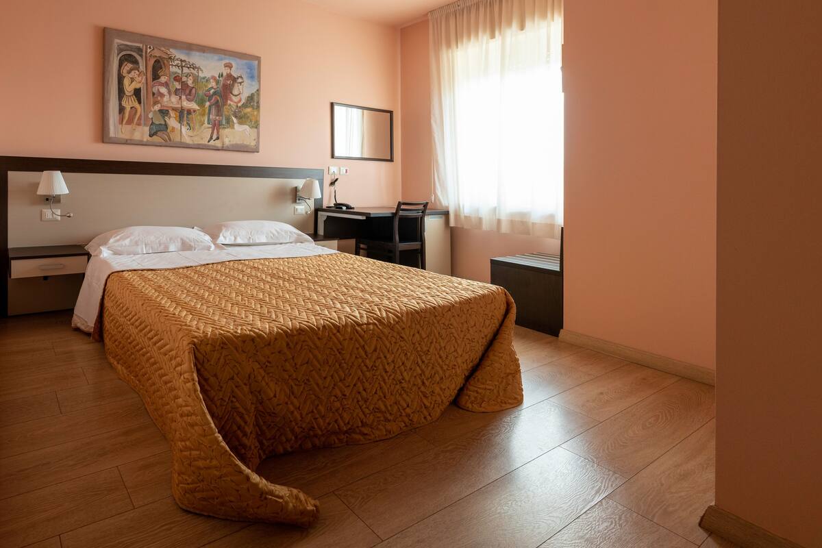 A hotel room with a large bed, a desk, a chair, a nightstand, a lamp, a mirror, and a painting on the wall.