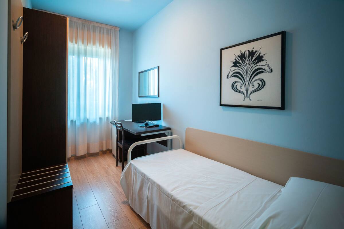 A hotel room with a bed, a desk with a computer, a chair, a wardrobe, and a framed artwork on the wall.