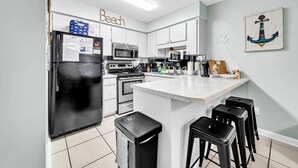 A modern kitchen with white cabinets, a black refrigerator, a microwave, an oven, and a sink. There are three black stools at the island. - PCB Beach Retreat~Steps to Beach, Pool & Amenities (Panama City Beach)