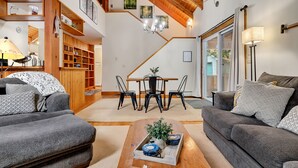 A cozy living room with a sofa, a coffee table with a tray, a dining area with a table and chairs, and a staircase leading to an upper level. - Great Sacandaga Waterfront Retreat (Day)