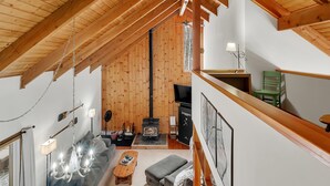 A cozy loft-style room with a high ceiling, wooden beams, a fireplace, a sofa, a coffee table, and a television. - Great Sacandaga Waterfront Retreat (Day)