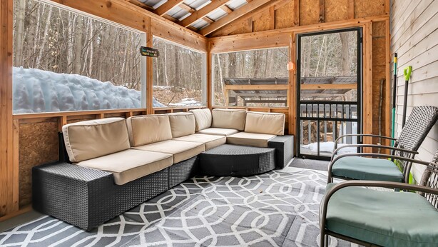 A cozy wooden cabin with a sectional sofa, a patterned rug, and a view of a snowy forest through large windows. - Great Sacandaga Waterfront Retreat (Day)