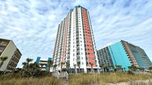 A tall residential building with multiple balconies, surrounded by other buildings and palm trees. - FANTASTIC OCEAN VIEWS FROM A BEAUTIFUL OCEAN FRONT CONDO! (Myrtle Beach)