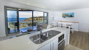 A modern kitchen with a large island, a built-in oven, and a sink. There is a dining area with white chairs and a table. A large window offers a view of the ocean and hills. - Shearwaters Appartment (Penneshaw)