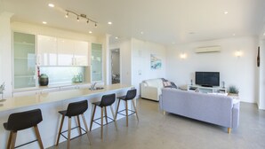 A modern kitchen with white cabinets, a central island with bar stools, a dining area with a sofa, and a TV. - Shearwaters Appartment (Penneshaw)