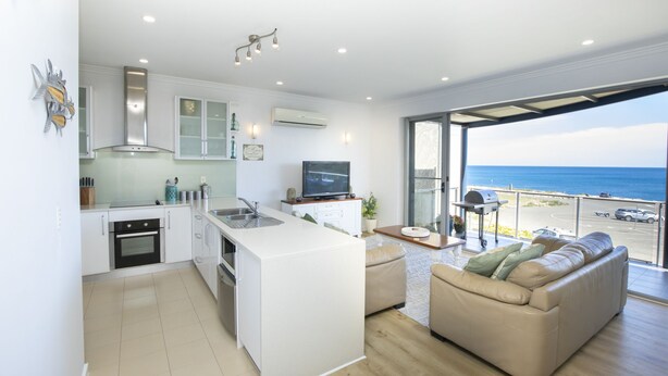 A modern kitchen with a central island, a dining area with a sofa, and a view of the ocean. - Shearwaters Appartment (Penneshaw)