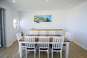 A dining area with a wooden table and white chairs, a painting above the table, and a window with a view of greenery. - Shearwaters Appartment (Penneshaw)