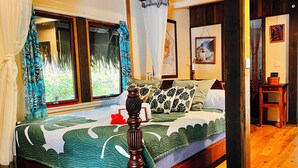 A wooden-framed bed with a green and white patterned bedspread, a decorative vase, and a view of greenery through a window with a curtain. - Orama Lodge Bora Bora - Le Fare  (Bora-Bora)