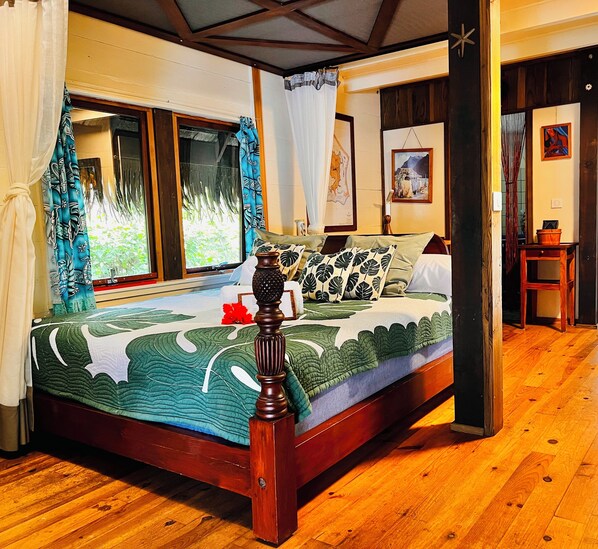 A wooden-framed bed with a green and white patterned bedspread, a decorative vase, and a view of greenery through a window with a curtain. - Orama Lodge Bora Bora - Le Fare  (Bora-Bora)