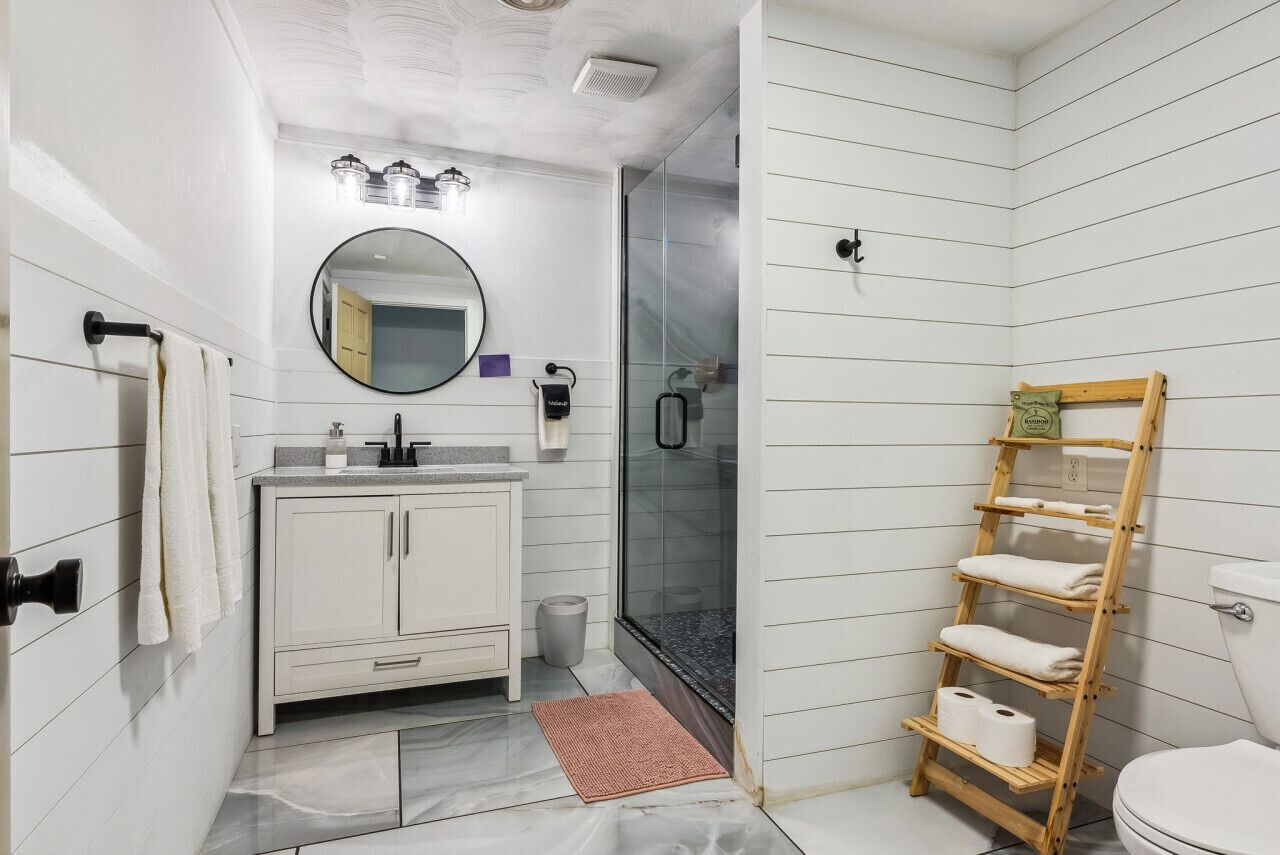Un baño con tocador blanco, un espejo redondo, cerramiento de ducha de vidrio y un perchero de madera.