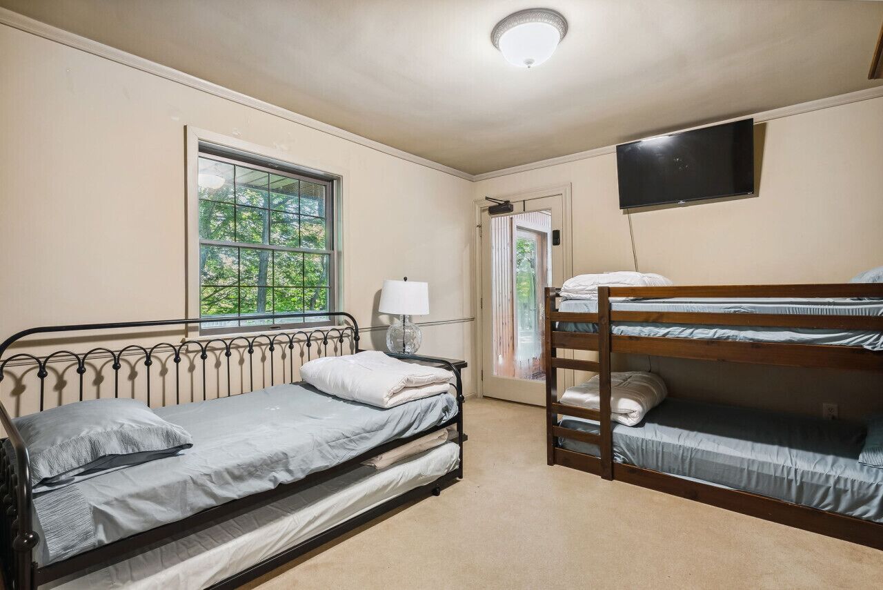 A room with a single bed, a bunk bed, a window with a view of trees, a flat-screen TV mounted on the wall, and a door leading outside.