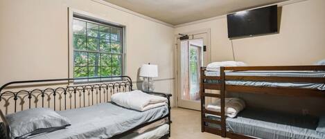 A room with a single bed, a bunk bed, a window with a view of trees, a flat-screen TV mounted on the wall, and a door leading outside.